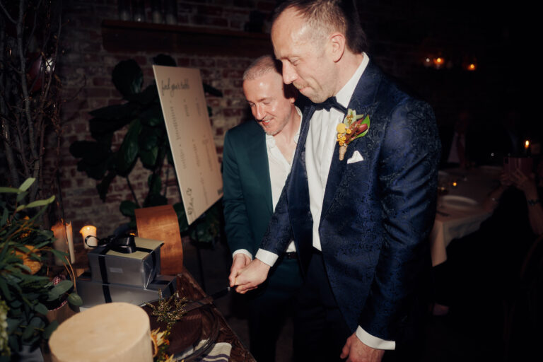 Two men in navy patterned tuxedos celebrate while cutting a cake at a dimly lit brick-walled venue, one with a boutonniere, candles flicker nearby
