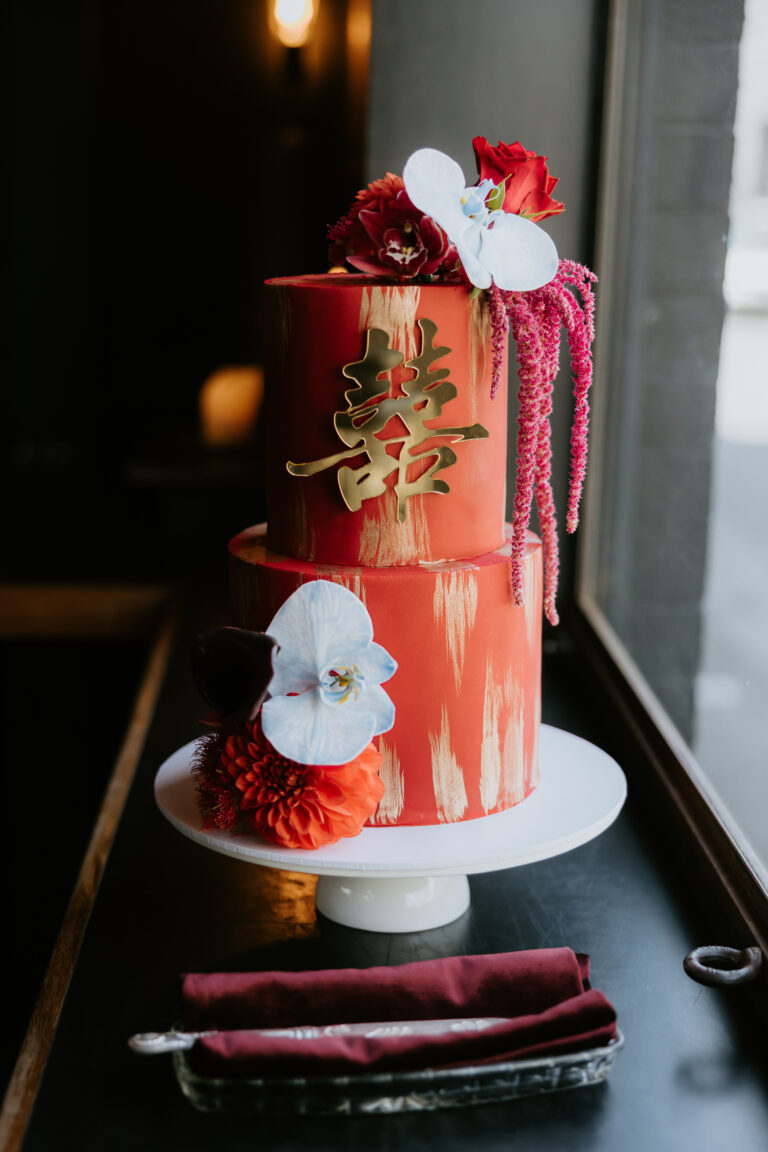 Red two-tier cake with gold kanji symbol, cascading pink and red flowers, white orchid, and a white cake stand, photographed by Dan Brannan