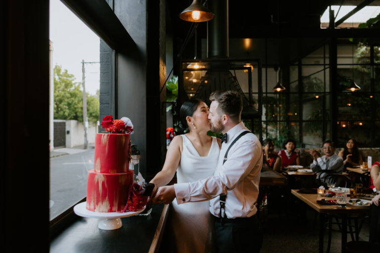 Bride in a white gown and groom in suspenders kiss beside a tall red drip wedding cake with flowers, inside a modern industrial venue with guests at tables