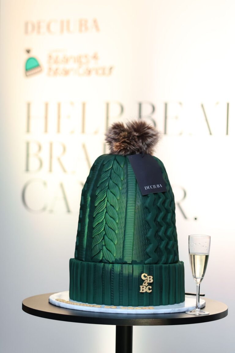 Diprose Media green knitted beanie cake with a fur pom-pom atop, textured cable-knit pattern, on a display table beside a champagne glass