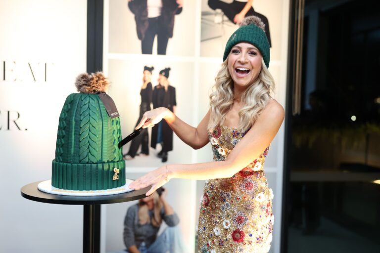 Smiling woman wearing a green knit beanie and a sparkling dress stands beside a tall emerald green tiered cake on a round black table, backdrop with fashion imagery
