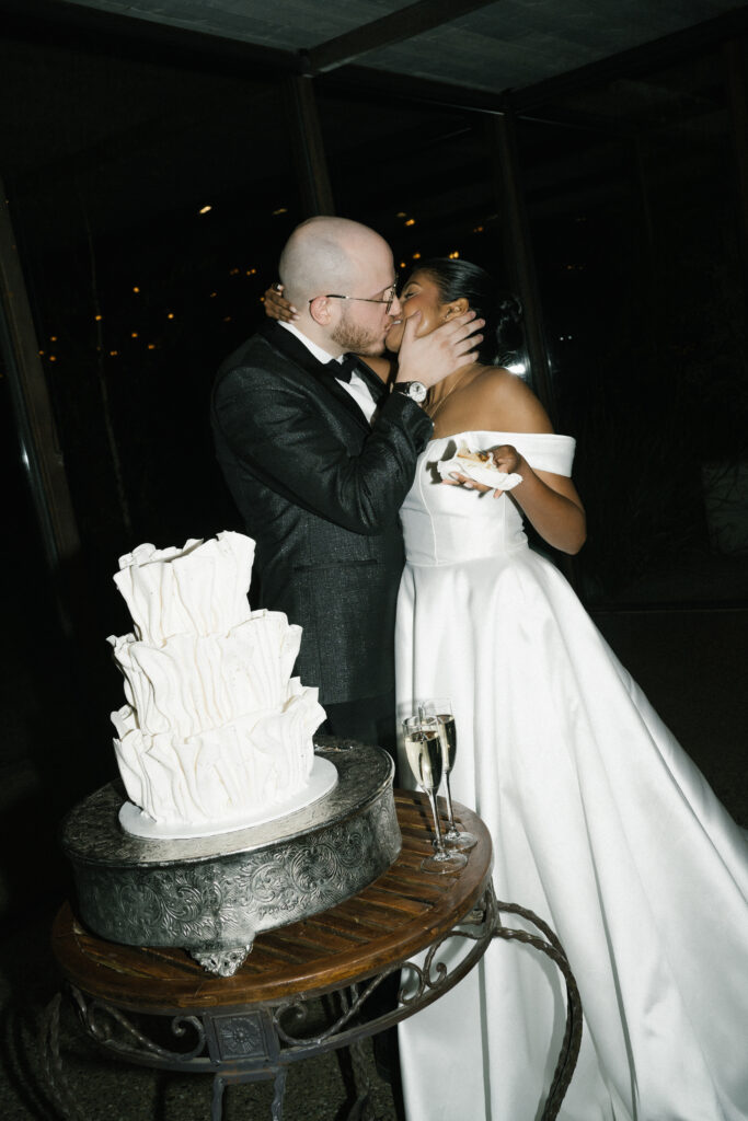 Ellie Rose McLean kissing her partner beside a white tiered cake; bride in an off-shoulder gown and groom in a dark suit, champagne nearby