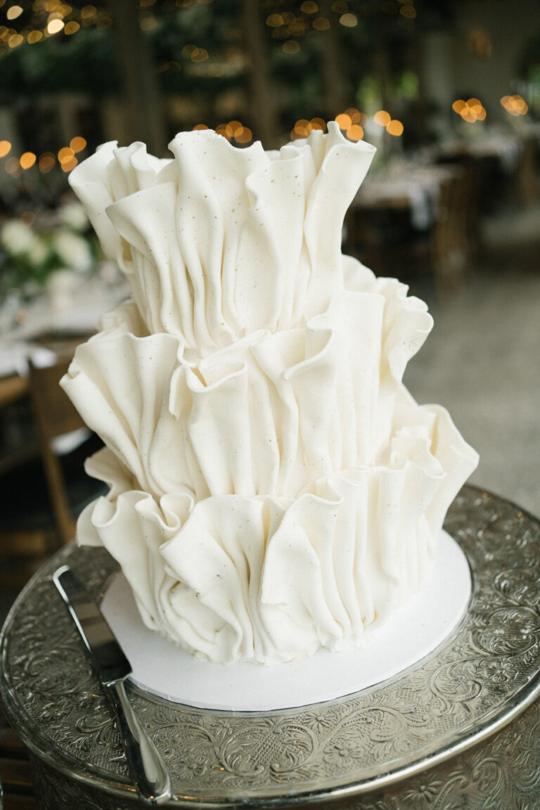 White ruffled cake by Ellie Rose McLean on an ornate silver stand, with a softly blurred warm background and delicate texture details
