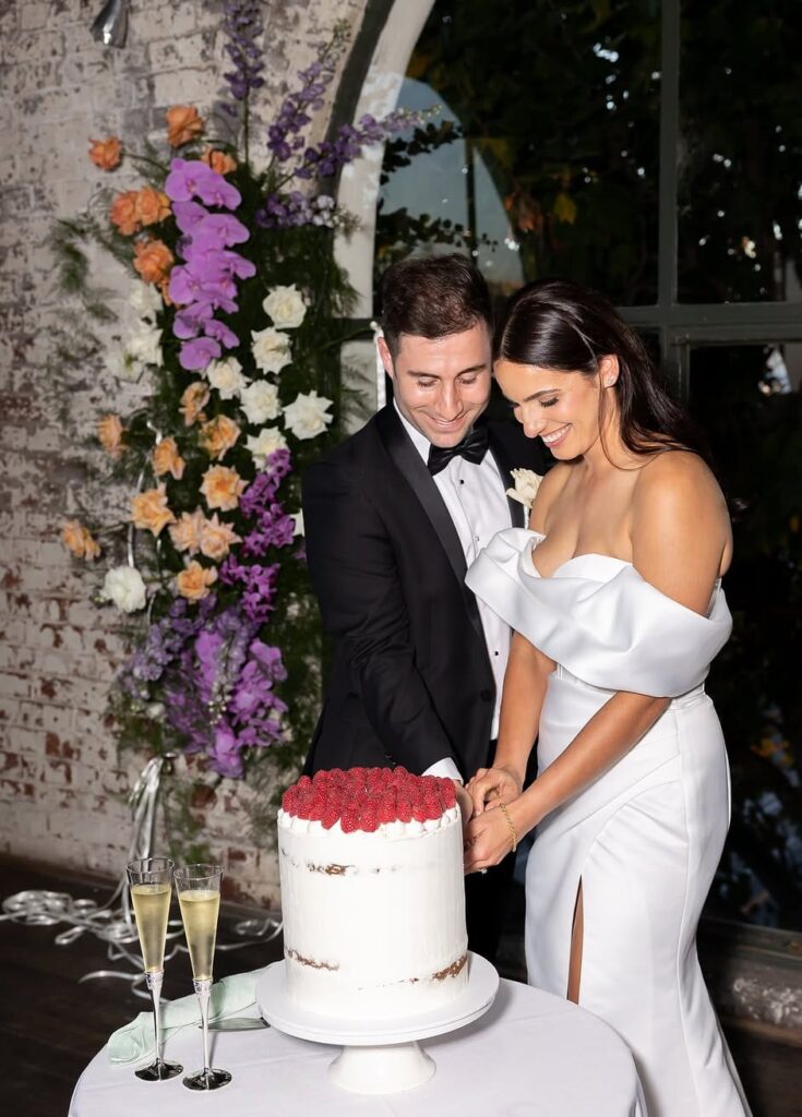 Bride in a white off-shoulder gown and groom in a black tuxedo cut a semi-naked cake topped with red berries, surrounded by a lush floral arch backdrop