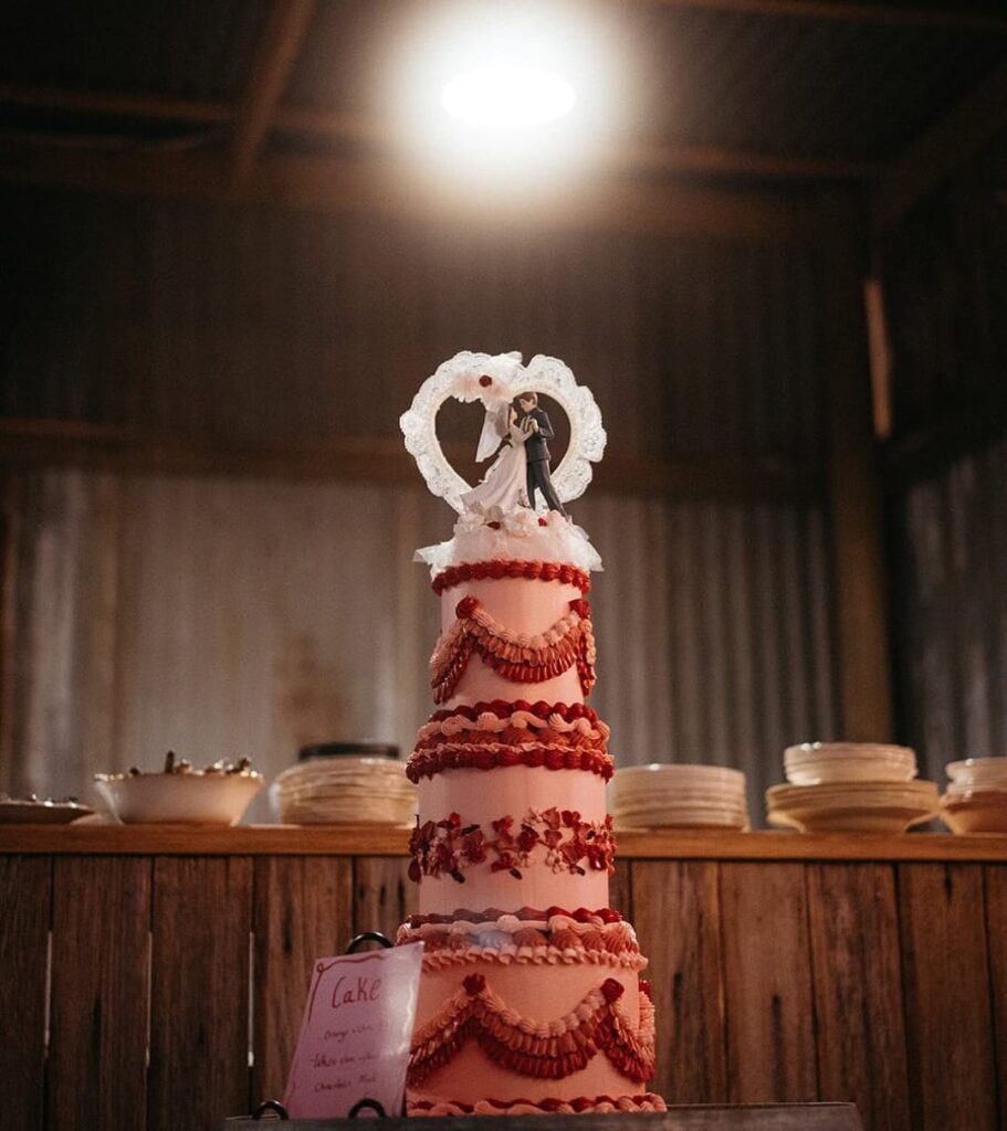 Elegant pink multi-tiered wedding cake with red piping, lace heart topper, and tiny bride and groom figurines set against a rustic backdrop