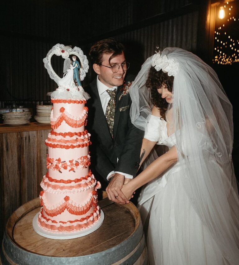 Pink tiered wedding cake with red ruffle icing sits on a wooden barrel while a bride in a white gown and veil and a groom in a dark suit hold hands nearby, warm lights in the background