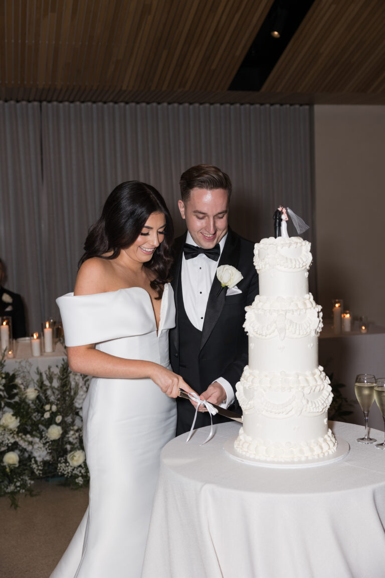 Studio Danielle Photography captures a bride in a white off-shoulder gown and groom in a black tuxedo cut a tall white three-tier cake with ruffled details at a candlelit reception.