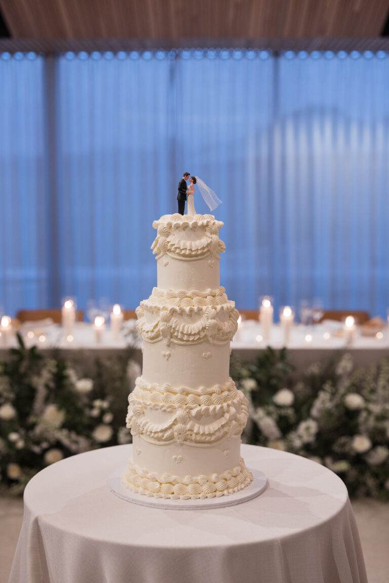 Studio Danielle wedding cake topper atop a multi-tier white cake with intricate cream frosting swirls and a bride and groom figurine