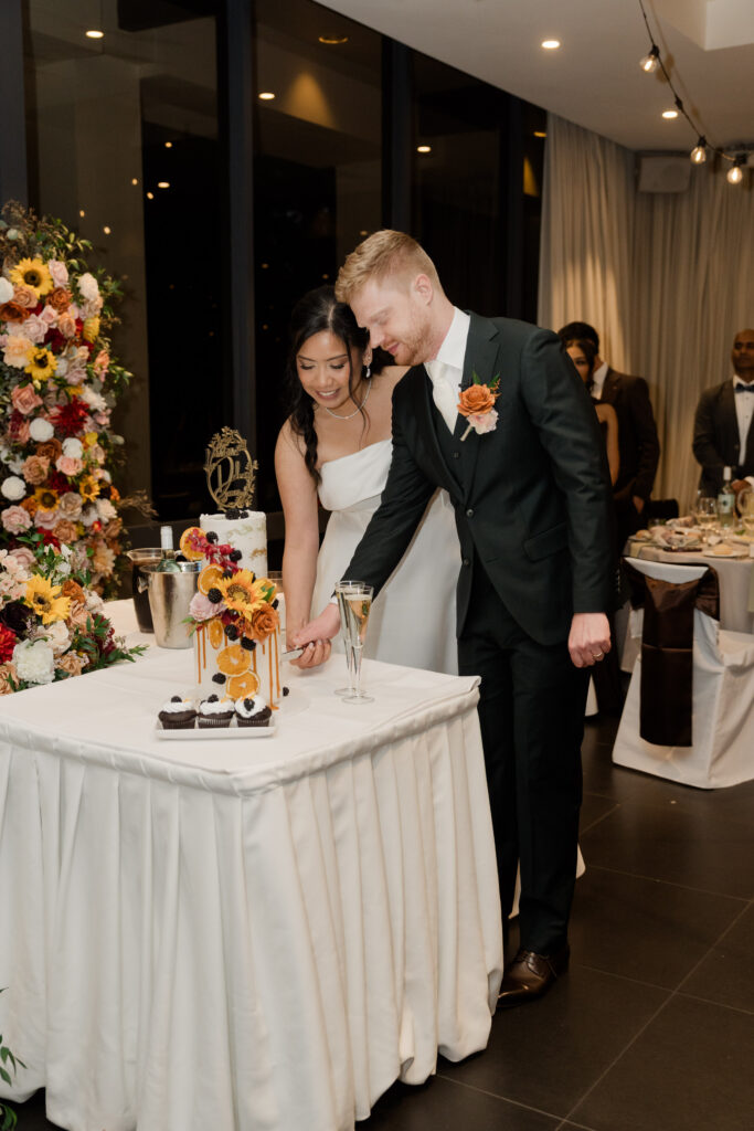 Bride and groom cutting a two-tier wedding cake decorated with orange slices, blackberries and colourful flowers.